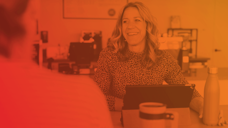 two women sit at a desk, one with an open laptop 