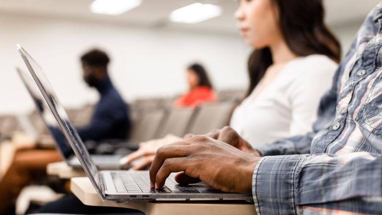 image of man in blue plaid shirt typing on keyboard. other students are blurry in the background