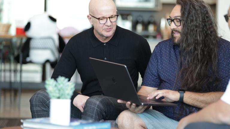 two men, one in a black shirt, the other in a blue shirt with long hair, sitting in front of a laptop working