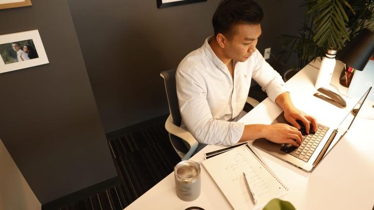 Man in a modern office space working on a laptop at a white desk, surrounded by a notebook, coffee cup, and desk lamp. The setting is clean, professional, and well-lit.