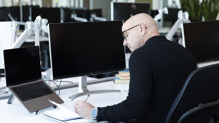 Bald man in a black sweater writing in a notebook while working at a desk with a laptop and multiple monitors in an open-concept office environment.