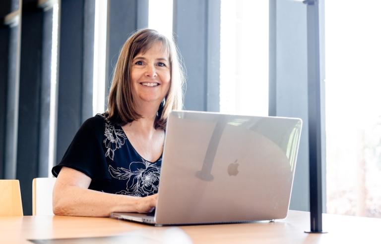 Woman smiles at camera while sitting at a desk with her laptop