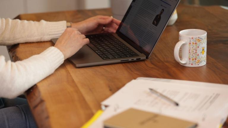 picture of a woman's hand on her laptop, with a notebook and mug on her desk.